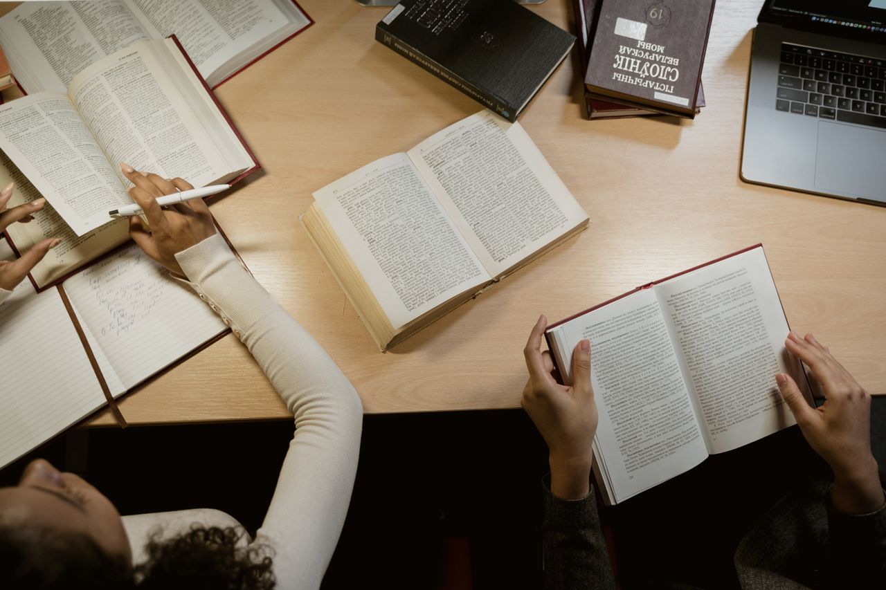 About Group of adults studying together with open books and laptops on a wooden table.